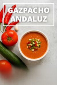 Gazpacho andaluz tradicional servido en bol blanco, decorado con trocitos de pepino, pimiento y tomate. Ingredientes frescos como tomate, pepino, ajo y pimientos rojos rodean el plato sobre fondo gris. Receta fácil y saludable típica de Andalucía